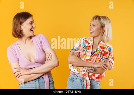 Portrait von zwei fröhlichen, lieblichen Freundinnen in Sommerkleidung, isoliert auf gelbem Hintergrund, Arme gefaltet Stockfoto