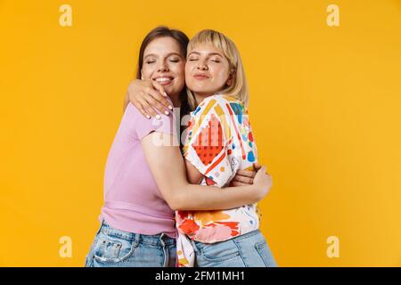 Portrait von zwei fröhlichen, lieblichen Freundinnen in Sommerkleidung, isoliert auf gelbem Hintergrund, umarmt Stockfoto