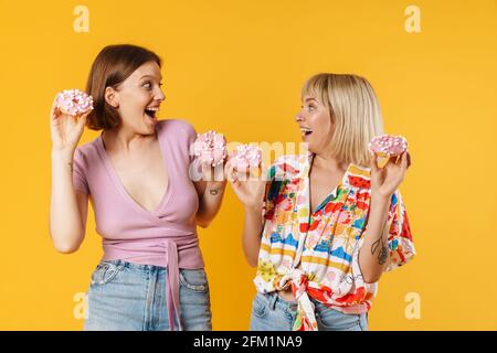 Porträt von zwei fröhlichen schönen Freundinnen tragen Sommerkleidung isoliert auf gelbem Hintergrund stehen, zeigt leckere Donuts, Spaß haben Stockfoto