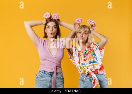 Porträt von zwei fröhlichen schönen Freundinnen tragen Sommerkleidung isoliert auf gelbem Hintergrund stehen, zeigt leckere Donuts, Spaß haben Stockfoto