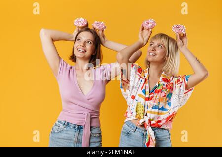 Porträt von zwei fröhlichen schönen Freundinnen tragen Sommerkleidung isoliert auf gelbem Hintergrund stehen, zeigt leckere Donuts, Spaß haben Stockfoto