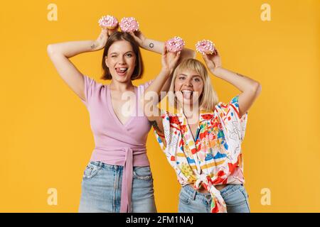 Porträt von zwei fröhlichen schönen Freundinnen tragen Sommerkleidung isoliert auf gelbem Hintergrund stehen, zeigt leckere Donuts, Spaß haben Stockfoto