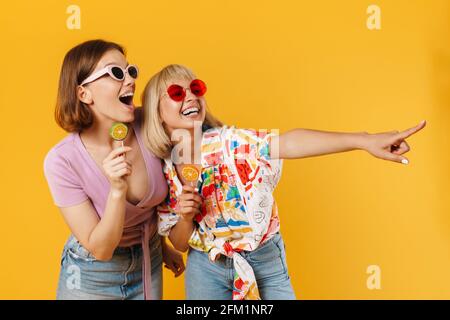 Portrait von zwei fröhlichen, lieblichen Freundinnen, die Sommerkleidung tragen und isoliert auf gelbem Hintergrund stehen, Lutscher halten und an Kopien herumschnappen Stockfoto