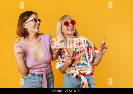 Portrait von zwei fröhlichen, lieblichen Freundinnen, die Sommerkleidung tragen und isoliert auf gelbem Hintergrund stehen, Lutscher halten und an Kopien herumschnappen Stockfoto