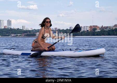 Glückliche schöne Frau mit Paddelbrett an einem Strand unter der Stadt. Sommer, Urlaub, SUP Paddleboarding oder Surfen, Reisen, Lifestyle-Konzept. Stockfoto