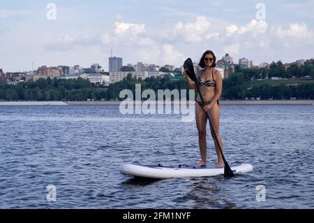 Glückliche schöne Frau mit Paddelbrett an einem Strand unter der Stadt. Sommer, Urlaub, SUP Paddleboarding oder Surfen, Reisen, Lifestyle-Konzept. Stockfoto