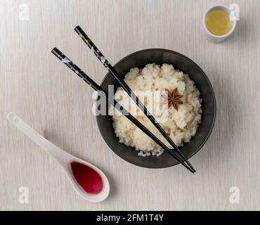 Ein Teller mit weißem Reis mit Sternanise auf einem Weiße Tischdecke mit orientalischen Essstäbchen oben und daneben Es ist ein Glas Sake und ein Löffel rot Soße Stockfoto