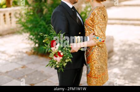Die Braut in einem ungewöhnlichen goldenen Kleid und der Bräutigam in einem Anzug umarmen sich, die Braut hält ihren Strauß in den Händen Stockfoto