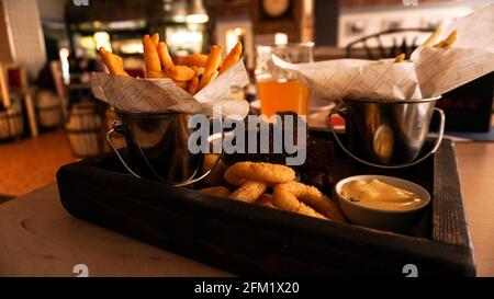 Bier-Set mit Snacks auf einem Holztablett. Zwiebelringe, Käsestäbe, Pommes in einem Eimer. Saucen. Snack für Freunde in einer Brasserie Stockfoto