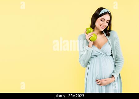 Freudige Schwangere hält frische Äpfel, während sie isoliert den Bauch berührt Auf gelb Stockfoto