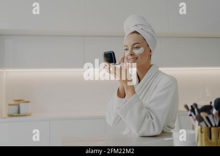 Glückliche Frau im weißen Bademantel mit Handtuch auf dem Kopf nehmen Pflege ihrer Haut nach der morgendlichen Dusche Stockfoto