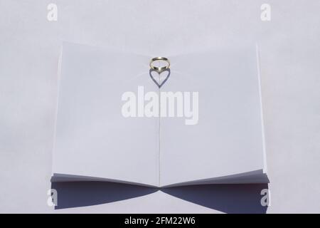 Sommerhochzeit-Mockup. Leeres Hochzeitsbuch auf einem weißen Tisch. Der Ehering wird auf das Buch gelegt und das Sonnenlicht erzeugt einen herzförmigen Schatten. Stockfoto