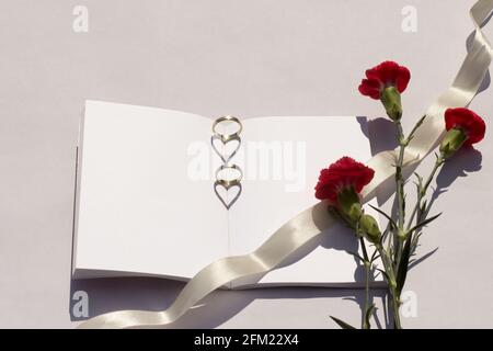 Sommerhochzeit-Mockup. Leere Hochzeitsbuch Blumen auf einem weißen Tisch. Eheringe sind auf dem Buch platziert das Sonnenlicht erzeugt einen herzförmigen Schatten Stockfoto