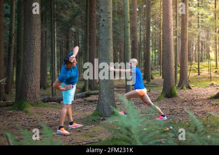 Paar macht Aufwärmübungen im Wald. Ein Mann, der Schultern und Rücken streckt, eine Frau lehnt sich an den Baum und macht Wadenübungen Stockfoto