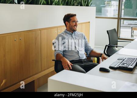 Junger Mann, der am Schreibtisch im Coworking Space sitzt. Freiberuflicher Mitarbeiter, der im Büro sitzt und lächelt. Stockfoto