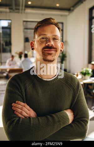 Selbstbewusster Geschäftsmann, der mit gekreuzten Armen lächelt und im Co-Working-Bereich steht. Glücklicher Manager, der die Kamera ansah und im Büro stand. Stockfoto