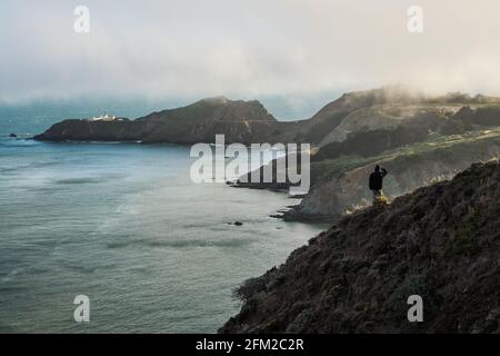 Mann in Outdoor-Tüchern, der alleine auf einem Hügel am Meer steht und den Point Bonita Leuchtturm an einem nebligen Tag in San Francisco überblickt, California, United S. Stockfoto