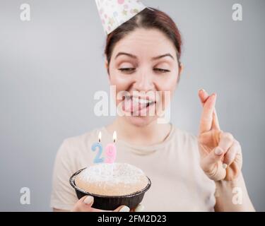 Glückliche kaukasische Frau, die ihre Zunge herausstreckte und mit gekreuzten Fingern die Kerzen auf dem Kuchen ausbläst. Das Mädchen feiert ihren 29. Geburtstag Stockfoto