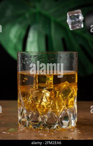 Nahaufnahme von Kristallglas mit Whiskey mit Eisflug, auf Holztisch und schwarzem Hintergrund mit grünem Blatt, vertikal Stockfoto