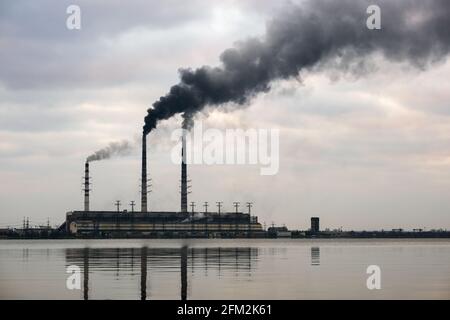Kohlekraftwerk hohe Rohre mit schwarzem Rauch, der nach oben die verschmutzende Atmosphäre mit den Reflexionen von ihm im Seewasser bewegt. Stockfoto