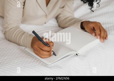 Zugeschnittenes Bild einer afroamerikanischen Frau, die im Bett liegt und Notizen in einem Tagebuch macht. Stockfoto