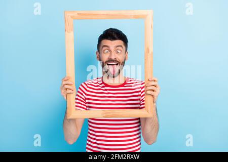 Foto von kindisch lustig jungen Kerl tragen rote Outfit halten Aussehender Holzrahmen mit Zunge isoliert blauen Hintergrund Stockfoto