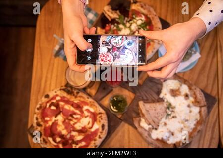 Weibliche Hände macht Fotos auf einem Smartphone-Tisch mit einer leckeren Pizza in einem Restaurant. Draufsicht Stockfoto