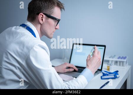Junger kaukasischer Arzt, der im Krankenhauslabor am Tisch arbeitet und eine Ampulle Medizin untersucht und Daten in den Computer eingibt. Neuer Impfstoff gegen Stockfoto