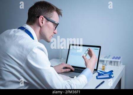 Junger kaukasischer Arzt, der im Krankenhauslabor am Tisch arbeitet und eine Ampulle Medizin untersucht und Daten in den Computer eingibt. Neuer Impfstoff gegen Stockfoto