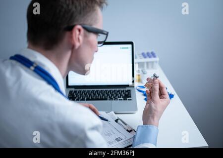 Junger kaukasischer Arzt, der im Krankenhauslabor am Tisch arbeitet und eine Ampulle Medizin untersucht und Daten in den Computer eingibt. Neuer Impfstoff gegen Stockfoto