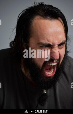 Porträt eines jungen Mannes, der schreit. Mann mit langen Haaren und langem schwarzen Bart brüllend, wütend. Verrückt nach dem Leben und frustriert. Stockfoto