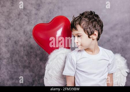 Junge mit Engelsflügeln versteckt hinter einem roten herzförmigen Ball. Stockfoto