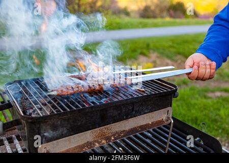 Beim Kochen köstlicher türkischer Adana Kebab auf einem tragbaren Holzkohlegrill an einem Picknickplatz kommt Rauch und Flamme aus dem brutzelnden Fleisch. Ein Mann ist an der Reihe Stockfoto