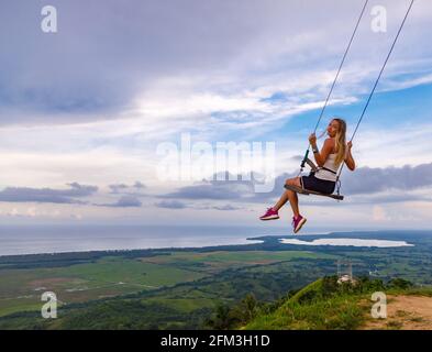 Ein junges Mädchen, blond, schwingt im Sommer auf einer Schaukel auf einem Berghang. Schwingt hoch in den Bergen über dem Tal. Dominikanische Republik. Untergehende Sonne Stockfoto