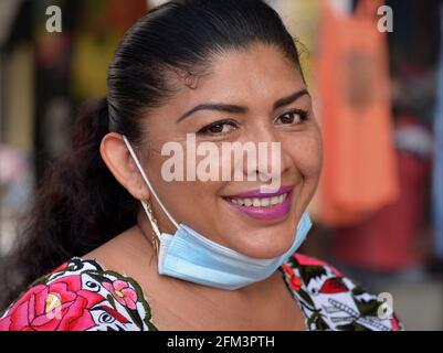 Eine Mexikanerin mittleren Alters mit schönen braunen Augen, rosa Lippenstift und blauer Gesichtsmaske unter ihrem Kinn lächelt während der globalen Corona-Virus-Pandemie. Stockfoto