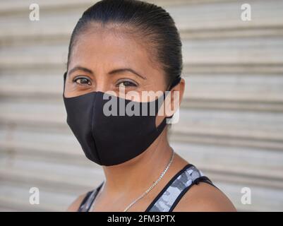 Eine Mexikanerin mittleren Alters mit schönen braunen Augen trägt während der anhaltenden weltweiten Coronavirus-Pandemie eine Gesichtsmaske aus nicht-medizinischem schwarzem Stoff. Stockfoto