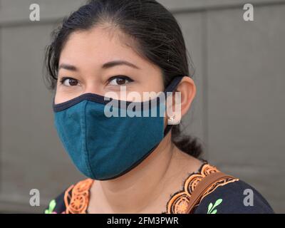 Die charmante, braun-äugige junge Mexikanerin trägt während der globalen Coronavirus-Pandemie eine blaue Gesichtsmaske aus nicht medizinischem Stoff und schaut in die Kamera. Stockfoto