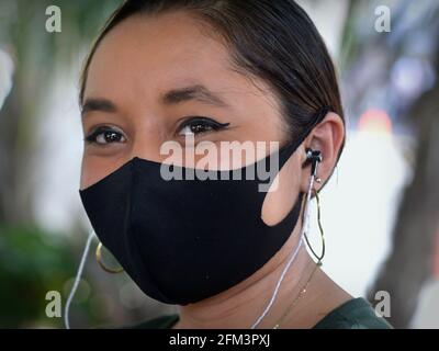 Die optimistische junge Mexikanerin mit Ohrhörern trägt während der Coronavirus-Pandemie eine schwarze Gesichtsmaske aus nichtmedizinischem Tuch und lächelt mit den Augen.