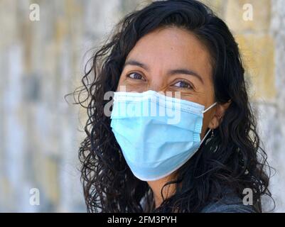 Positive optimistische kaukasische Frau mit nassen langen schwarzen Locken und lächelnden braunen Augen trägt während der Corona-Pandemie eine hellblaue chirurgische Gesichtsmaske. Stockfoto