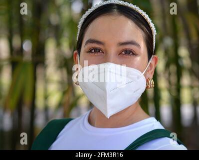 Die junge mexikanische brünette Latina-Frau lächelt mit ihren schönen braunen Augen und trägt während der globalen Coronavirus-Pandemie eine weiße KN95-Gesichtsmaske. Stockfoto