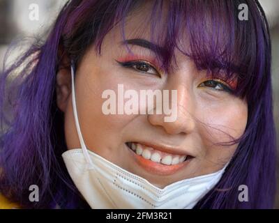 Junge glückliche indigene Südamerikanerin mit aufwändigem Augen-Make-up und blau gefärbtem Haar zieht ihre KN95-Gesichtsmaske herunter und lächelt für die Kamera. Stockfoto