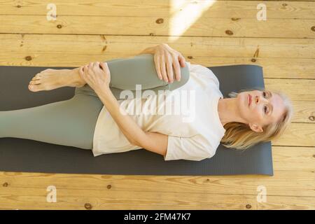 Morgengymnastik. Energische ältere Frau macht Stretching-Aufwärmübungen, während sie zu Hause auf der Yogamatte auf dem Boden liegt. Ältere Frau nach einem gesunden Lebensstil im Ruhestand Stockfoto