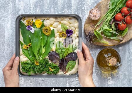 Weibliche Hände dekorieren rohe Blumenmalerei Focaccia, Garten Sauerteig Kunst mit saisonalen Bärlauch Blätter. Stockfoto