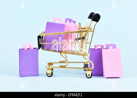 Goldener Einkaufswagen gefüllt mit pinken und violetten Papiereinkäufen Taschen auf blauem Hintergrund Stockfoto