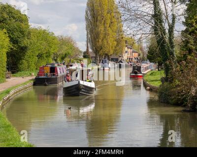 Canal and River Trust Trainingsboot, das an festgetäuten schmalen Booten auf dem Grand Union-Kanal, Stoke Bruerne, Großbritannien, vorbeifährt Stockfoto