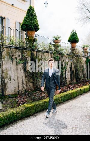 Der ernsthafte Bräutigam geht durch einen grünen Park vor dem Hintergrund eines Steinzauns und alter Gebäude. Comer See Stockfoto