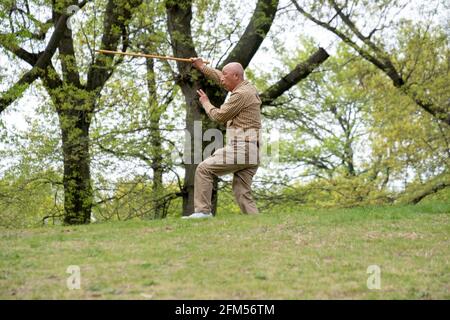 Ein älterer asiatischer Amerikaner, wahrscheinlich Chinese, macht Tai-Chi-Übungen mit einem langen Stock. Auf einem Hügel in einem Park in Queens, New York City. Stockfoto
