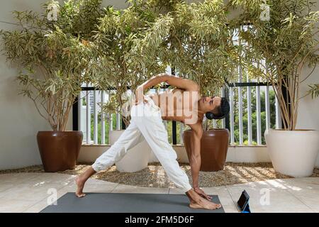 Lächelnder junger Brasilianer, der mit seinem Kopftelefon und seinen Hüften online Yoga-Kurs macht. Stockfoto