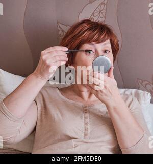 Ältere Frau mit kurzen roten Haaren in Hauskleidung Make-up zu tun. Sie sitzt auf dem Bett und malt ihre Wimpern mit einem Pinsel und Mascara, Blick in ein s Stockfoto