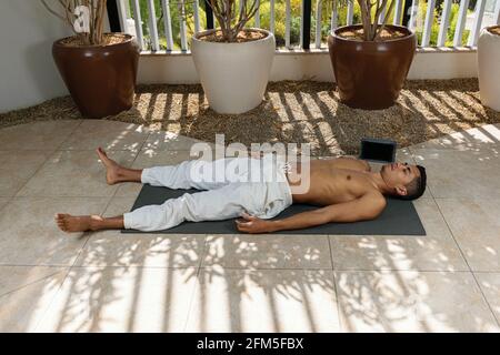 Junger brasilianischer Mann, der Online-Yoga-Kurs macht, in der Leichenstellung. Stockfoto